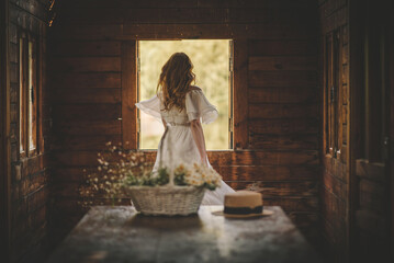 beautiful girl with red long curly hair whirls waving her hair near the window in an old wooden house in a white dress in the foreground a table on which a basket with flowers and a straw hat 