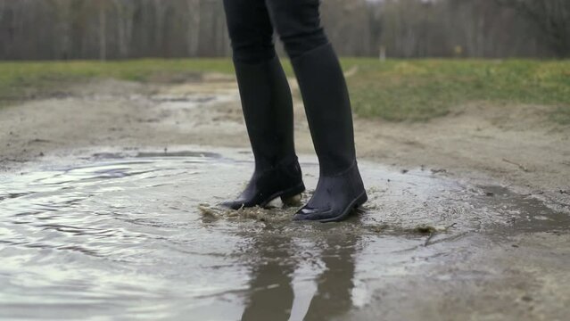 A woman in black rubber boots jumps up in a muddy puddle.