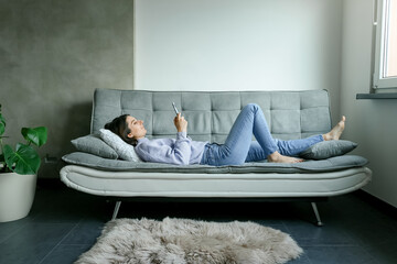 Back view of happy young woman in sweater laying on sofa checking her phone