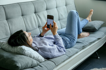 Back view of happy young woman in sweater laying on sofa checking her phone