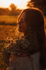 silhouette of a beautiful young girl with red curly hair in a summer dress standing with closed eyes and holding in his hands near the face of chamomile at sunset, side view close up