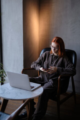a red-haired freelance woman with glasses works remotely on a laptop in a cafe.x