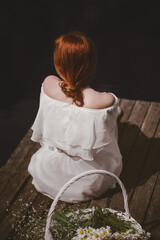 beautiful young girl with red hair gathered in a bundle in a white dress sitting on a wooden pier next to her stands a white straw basket with field rum rural scene on a sunny day rear view 