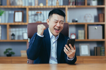 Successful Asian working in a classic office holding a phone, celebrating victory, gesture of...