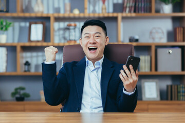 Successful Asian working in a classic office holding a phone, celebrating victory, gesture of...