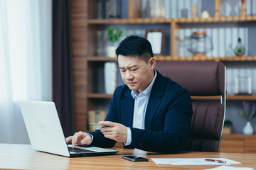 Upset Asian man, trying to make an online purchase, sitting in the office, uses a credit card, and a laptop, trying to remember the pincode