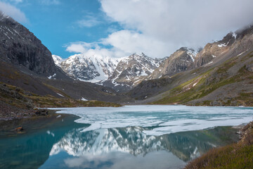 Fototapeta na wymiar Atmospheric landscape with frozen alpine lake and high snow mountains. Ice floats on transparent water surface of mountain lake. Awesome scenery with large snowy mountains reflection in clear lake.