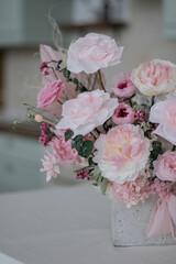Beautiful bouquet of peonies and pink roses on the table. In the background is the interior of a modern white kitchen. Concept of home comfort.