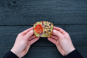 Hands holding avocado toast with grilled mushrooms and grilled tomatoes on a dark wooden table. Healthy food and vegan diet concepts