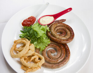 Grilled round-spiral grilled sausage with fried onions, green salad and sos in a red spoon on a white plate.