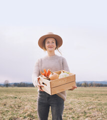 Woman with local organic food on agricultural field area. Customer or farmer with wooden box of vegetable at local farm market outdoors. Sustainable shopping without plastic. Small business concept.