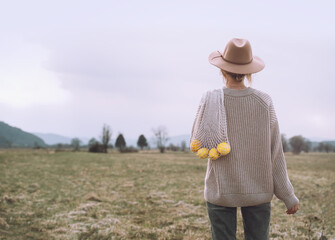 Creative image of young woman holding lemons in reusable cotton mesh bag in field. Hipster girl wearing minimalist style clothes walking in nature. Concept of slow fashion and life, eco lifestyle. - obrazy, fototapety, plakaty