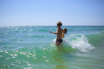 Summer happy family of six years blonde child playing and jumping water waves embracing woman mother in sea shore beach