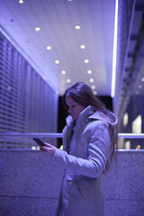 Young millennial woman using mobile phone taxi app or texting on social media standing near office modern architecture building with purple blue violet neon light at night