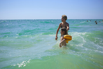 Summer happy family of six years blonde child playing and jumping water waves embracing woman mother in sea shore beach