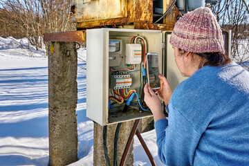 Taking readings from electricity meter using handy by woman in countryside outdoors. - obrazy, fototapety, plakaty