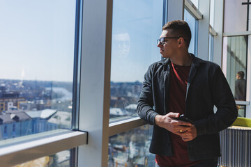A calm confident man in a fashionable outfit and glasses stands with a smartphone in his hands near the window and looks at the city from a skyscraper in the daylight