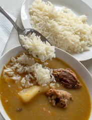 A white bowl of Dominican sancocho with yucca, chicken, pork, squash and more, served with white rice and a metal spoon.