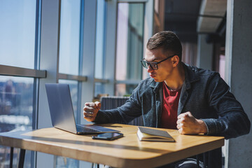 Portrait of a man, IT professional, working remotely with a modern laptop, sitting at a table and smiling at the camera during a break, a happy human programmer in vision correction glasses