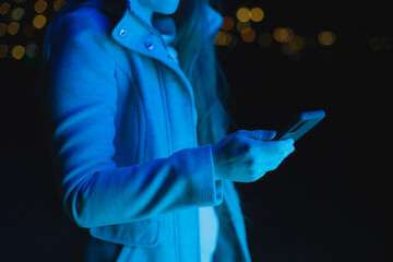 young woman holding mobile phone, texting and using social media in blue neon light in city at...