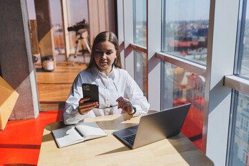 Young business woman in glasses speaks on the phone in the office. Business woman texting on the phone and working on a laptop. A young business woman is sitting at a table in a cafe. Smiling woman.