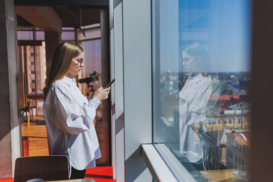 Smiling woman in glasses in casual wear watching a positive video on a phone connected to wi-fi, a female blogger in a white shirt against a large window, enjoying relaxation during remote work.