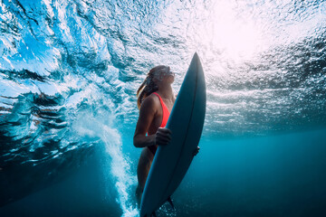 Surfer girl with surfboard underwater with ocean wave and sun light.