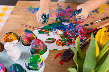Fun and handmade craft of little child during painting ecolorful easter eggs. Hands of kid and seasonal creativity. Yellow tulips and decoration for spring holiday. Close up. 