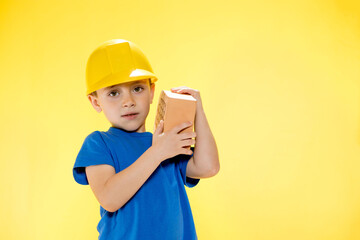 Boy in a construction helmet holds a brick in his hands on yellow background