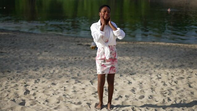 Joyful African American young barefoot woman in dress dancing in slow motion on sunny sandy beach outdoors. Wide shot portrait of happy cheerful beautiful lady having fun enjoying summer leisure