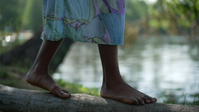 Slim African American female feet walking on tree trunk on river bank. Unrecognizable young barefoot woman in dress strolling outdoors in summer spring park enjoying leisure. Slow motion
