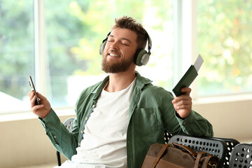 Handsome man with passport, mobile phone and headphones listening music in hall of airport