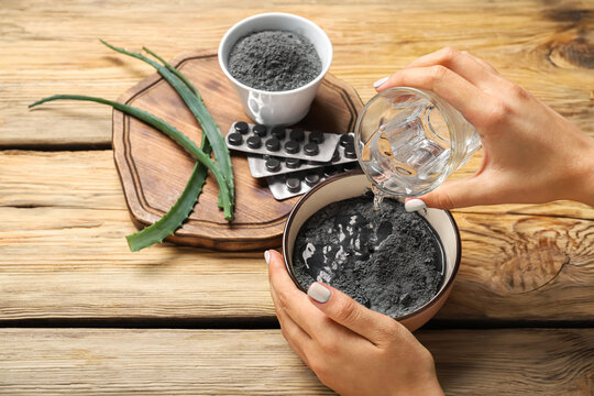 Woman pouring water from glass into bowl with activated carbon facial mask on wooden background, closeup - Powered by Adobe