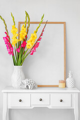 Vase with beautiful gladiolus flowers and blank poster on chest of drawers near white wall