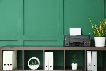 Shelf unit with modern printer, houseplant and folders near green wall