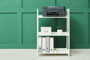 Shelf unit with modern printer, houseplant and folders near green wall in room