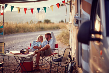 Couple in love, sitting and smilling at each other in front of camper rv. Fun, travel, togetherness, lifestyle, love concept.
