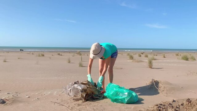 Mature woman with white hat and green gloves folding a large nylon picked up on the beach and put it in a garbage bag. 