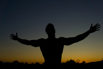 Silhouette of muscular man posing shows his muscles against the sky at sunset. 