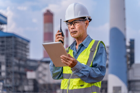 Senior supervisor engineer in his work cloths with white hard hat, safety glasses using his walkie talkie radio to communicate with his team at the power plant site