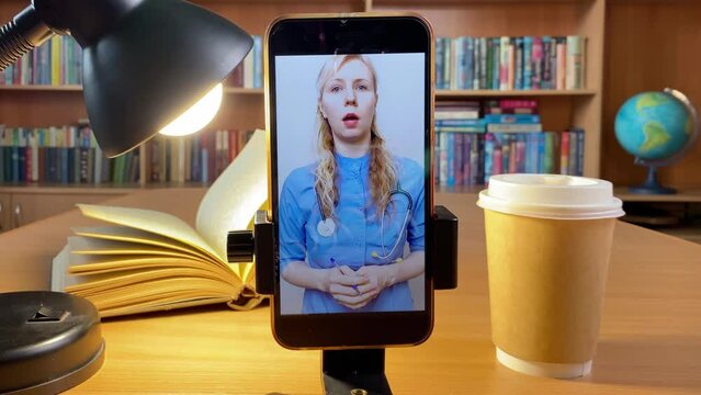 Online education. Video Conference Call in library. Young woman in blue uniform and stethoscope makes explanations. Doctor gives consultation online. Books, shelves and globe on background