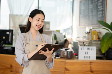 Pretty Asian barista in apron holding a clipboard and standing in front of a coffee shop counter. Startup concept of business owner.