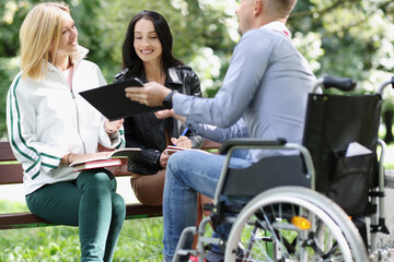 Two women and a man in a wheelchair look at tablet while sitting in park