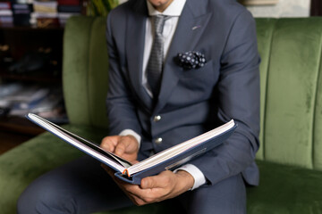 Man in expensive tailored suit sitting on sofa, choosing material from catalogue in studio.