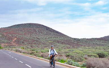 Senior adult woman wearing helmet and eyeglasses cycling in outdoors with backpack on shoulders. Cloudy sky and mountain in background