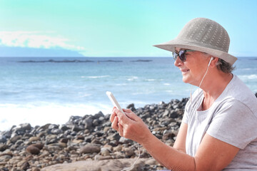 Smiling senior woman wearing hat and sunglasses sitting on the beach using mobile phone sending message. Holding telephone with white cover . Horizon over water