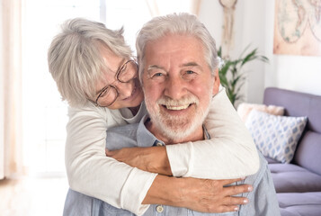 Fototapeta na wymiar Beautiful senior caucasian woman lovingly hugging her seated husband. Smiling elderly couple white haired looking at camera