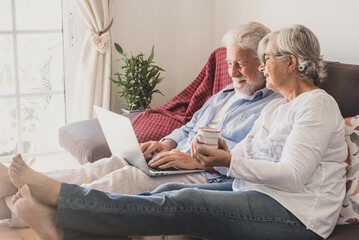 Senior caucasian couple sitting at home on sofa using laptop computer. Two elderly retirees enjoying free time and technological devices