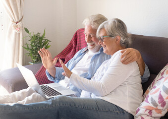 Happy caucasian senior couple sitting at home on sofa using laptop in video call waving hands. Two elderly retirees enjoying free time and technological devices