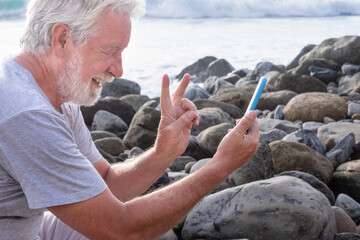 Smiling senior bearded man having video conversation with mobile phone while sitting on the rock beach facing the sea